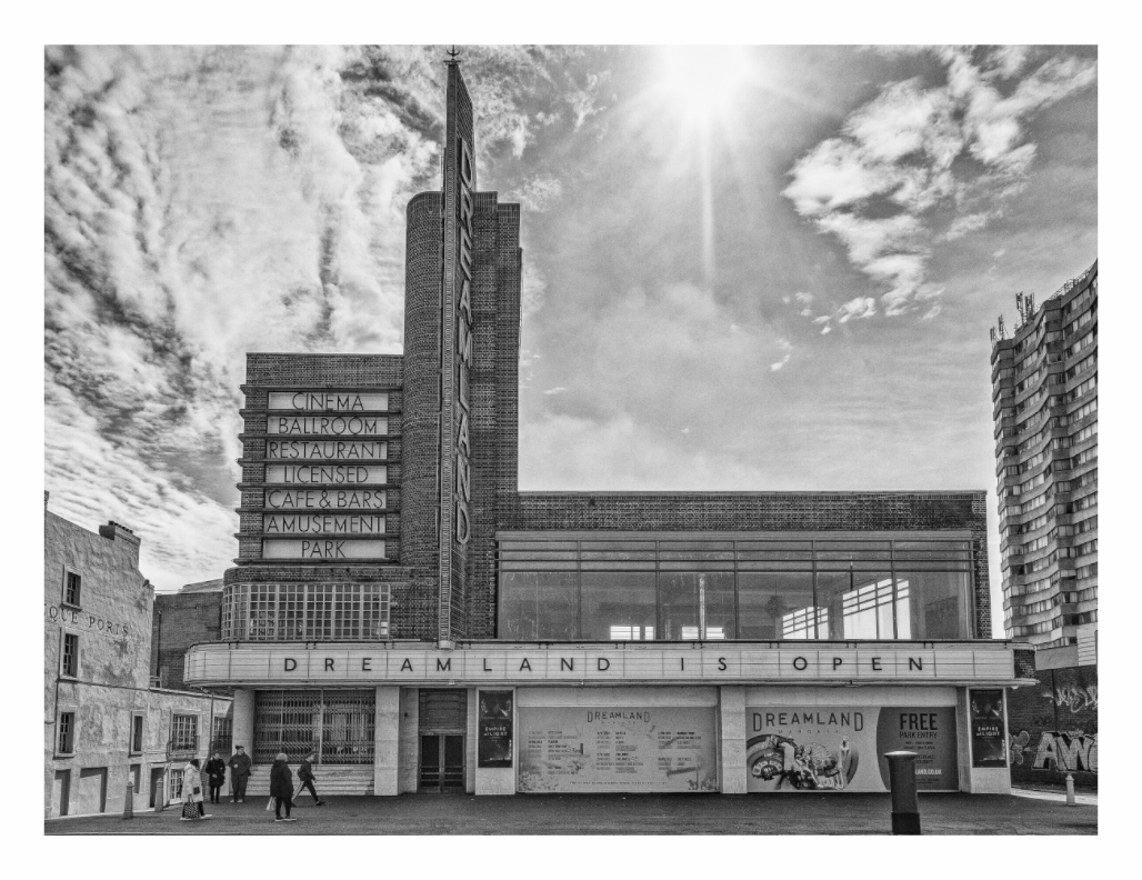 Black and white image of a cinema with 'Dreamland is Open' sign against a cloudy sky, Monochrome street photo capturing the exterior facade of the historic Dreamland amusement park building in Margate.