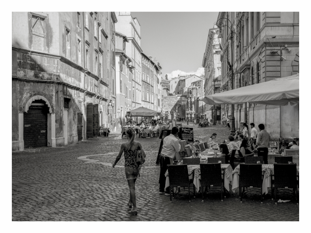 Black and white street scene with people and outdoor seating area. Black and white fine art print capturing a street scene in a Roman Piazza, featuring historical architecture, cobblestones, and figures walking.