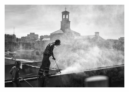Black and white fine art photo of a boat worker in Ramsgate, Kent, jetwashing rust from the hull of a large, industrial barge on a dry slip.