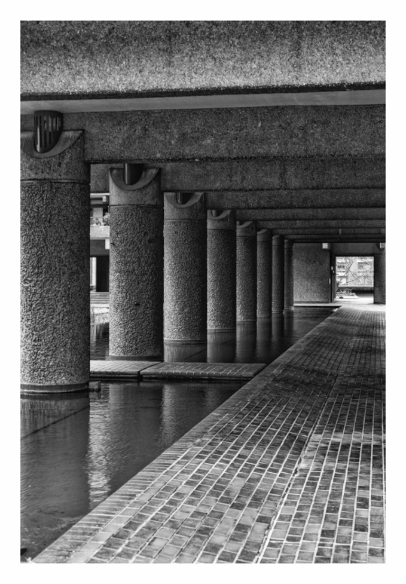 Concrete columns and a brick pathway in an urban setting, Black and white fine art photograph of the Barbican Centre in London, highlighting its Brutalist concrete architecture.