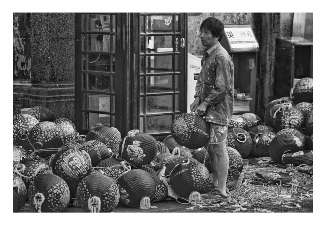 Person standing among large spherical containers in a market setting, Black and white fine art street photograph of a bustling China Town street in London, possibly showing New Year decorations.