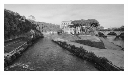 Black and white photograph of a river with a building and trees on the bank, and a bridge in the distance, Monochrome fine art print showing the facade of the iconic Fatebenefratelli Hospital building on Tiber Island, Rome.
