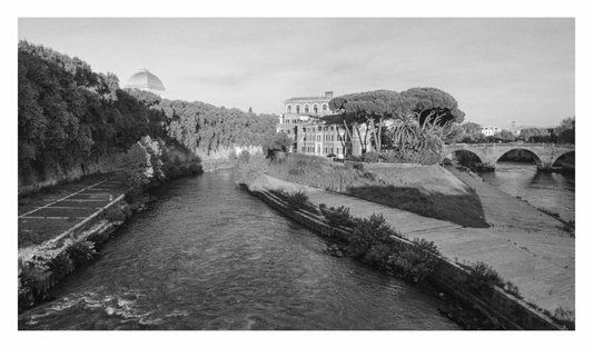 Black and white photograph of a river with a building and trees on the bank, and a bridge in the distance, Monochrome fine art print showing the facade of the iconic Fatebenefratelli Hospital building on Tiber Island, Rome.