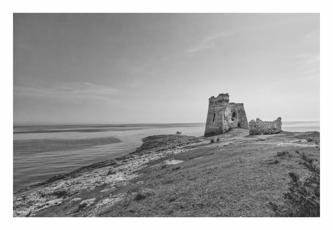Black and white photograph of an old castle ruin on a hill overlooking a coastal landscape. Black and white architectural print of a ruined, ancient coastal fortification or fort wall in Puglia, Italy.