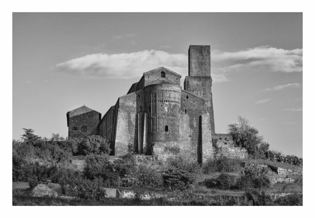 Black and white photograph of a historic stone building with a tower, surrounded by trees and clear sky. Black and white fine art photo of the Romanesque facade of the San Pietro Church in Tuscania, Italy, featuring an intricate rose window and carvings.