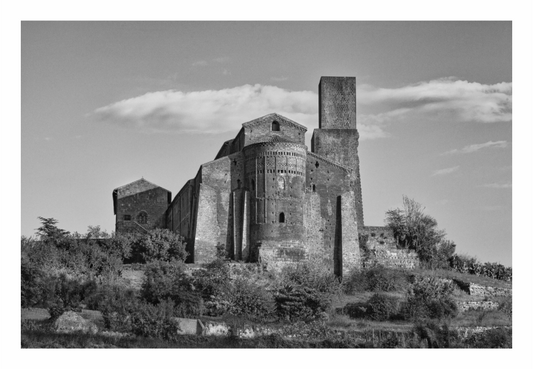 Black and white photograph of a historic stone building with a tower, surrounded by trees and clear sky. Black and white fine art photo of the Romanesque facade of the San Pietro Church in Tuscania, Italy, featuring an intricate rose window and carvings.