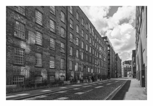 Black and white street scene with brick buildings and a cloudy sky. Black and white fine art photo of a narrow, high-contrast Manchester street scene, using linear perspective to show deep urban shadow.