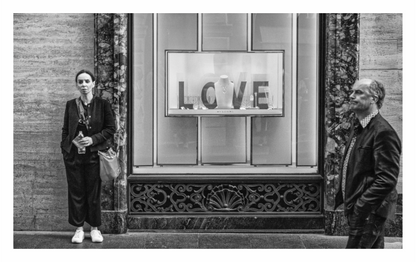 Two people standing in front of a store window with 'LOVE' displayed, Black and white fine art street photograph capturing a human interaction on Via dei Condotti, Rome's luxury shopping street. 