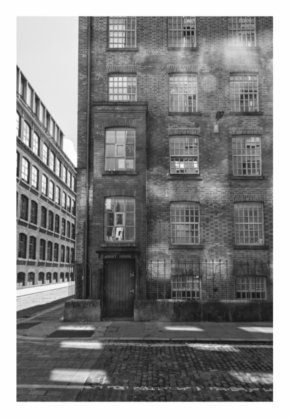 Black and white photograph of a brick building with windows on an urban street, Black and white fine art photograph of a historic cobbled street in Manchester with highly textured pavement and strong linear perspective.