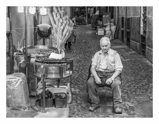 Man sitting on a chair next to a small stove with pots on a cobblestone street, Black and white fine art street portrait of a Hot Chestnut vendor operating a cart at the Spanish Steps in Rome, Italy.