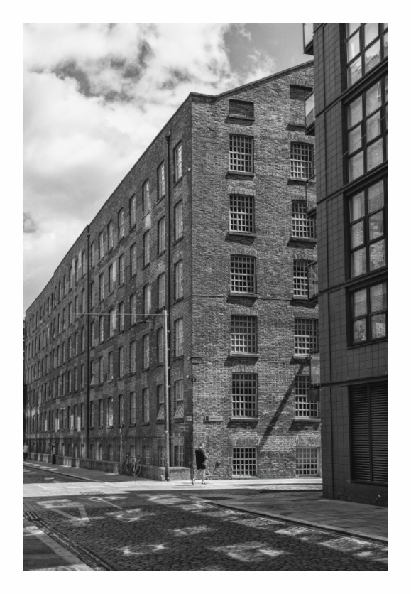 Black and white photograph of a large brick building with modern glass facade. Black and white fine art photo of a historic cobbled street in Manchester, utilizing high contrast to emphasize the pavement texture and shadows
