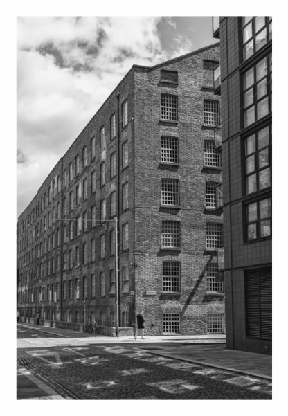 Black and white photograph of a large brick building with modern glass facade. Black and white fine art photo of a historic cobbled street in Manchester, utilizing high contrast to emphasize the pavement texture and shadows