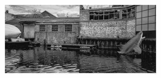 Black and white image of a building with a large shark model in front of it, Black and white street photography of a "Shark Infested" warning sign next to a London canal or waterway.