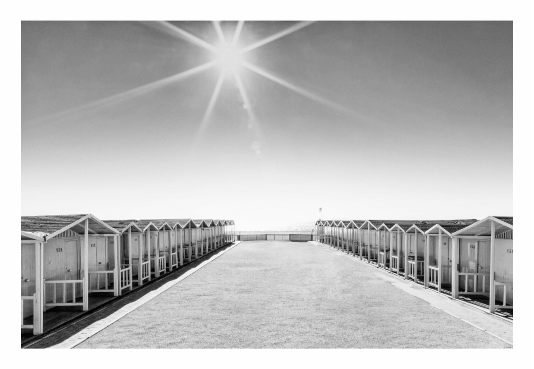 Black and white photo of a sunlit beach with rows of small huts. Black and white fine art photo of a row of traditional beach huts on the coast of Ostia, Lazio, Italy, highlighting their repetitive forms.