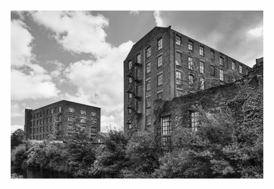 Two brick buildings on a canal with trees and a cloudy sky, Black and white fine art photo of a historic brick canal warehouse in Manchester, featuring the building reflected in the water.