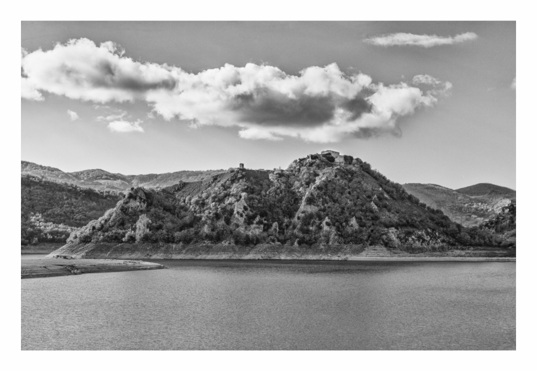 Black and white landscape of a lake with a hill and trees under a cloudy sky. Black and white landscape print of Castel di Tora village perched on a hill overlooking the clear waters of Lake Turano in Lazio, Italy.