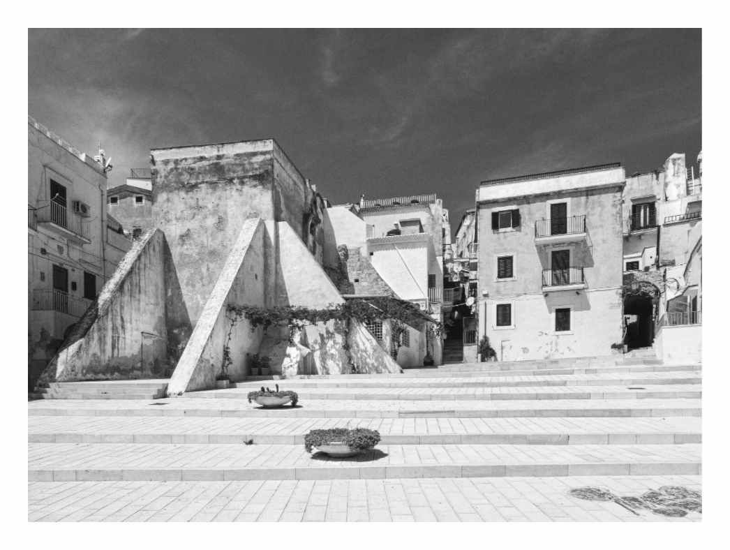 Black and white photo of a square with architectural structures and steps. Black and white street photo of a quiet, historic town square in Puglia, Italy, capturing the traditional architecture and paved area.