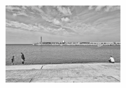 Black and white scene of people by a waterfront with a lighthouse in the distance. Black and white architectural photo of wide, descending Victorian steps near the harbor arm in Margate, UK.