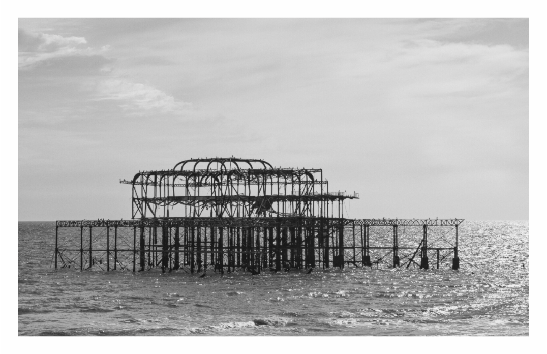 Black and white photograph of an old pier structure in the middle of a body of water, Dramatic monochrome print capturing the iconic, derelict West Pier silhouette against the water.