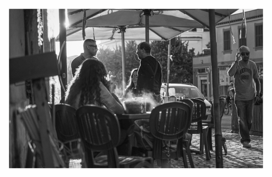 People sitting at outdoor tables under umbrellas on a street, Monochrome architectural print capturing a deserted, sun-drenched, narrow alley in an Italian town.