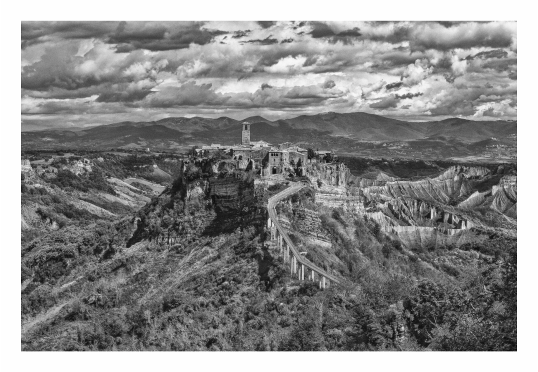 Black and white landscape of a castle on a cliff with a cloudy sky. Monochrome print of the "dying city" of Bagnoreggio, showing the dramatic hilltop architecture and the vast, barren valley beneath it.