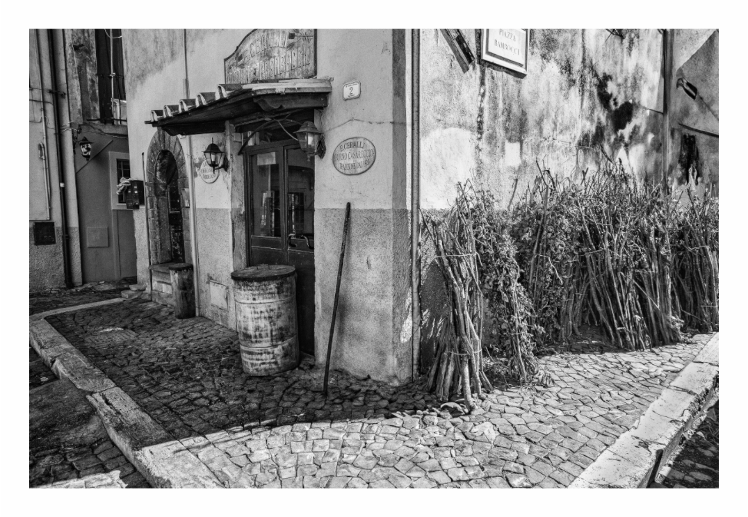 Black and white street scene with building entrance and barrels on a cobblestone street, Monochrome street photography print showing a historic stone-paved corner with European architecture in Italy.