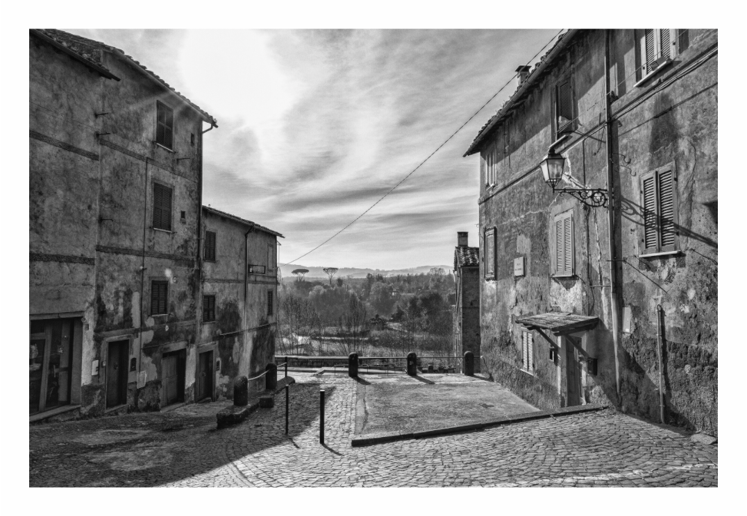 Black and white fine art photograph of a narrow Italian street, emphasizing the high-contrast lighting and linear perspective of the architecture.