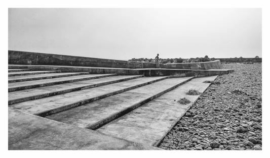 Black and white photograph of a concrete walkway with steps leading into a field. Monochrome fine art print capturing a solitary figure on geometric beach steps, emphasizing contrast and solitude.