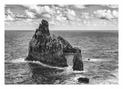 Black and white photograph of a rocky island in the ocean with clouds above. Monochrome print of the Madeira coastline, showing powerful ocean waves crashing against a massive, dark seaside cliff.