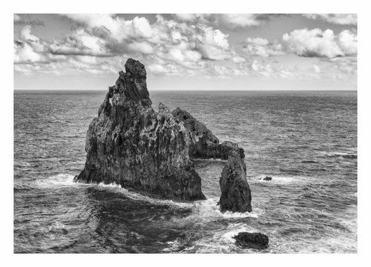 Black and white photograph of a rocky island in the ocean with clouds above. Monochrome print of the Madeira coastline, showing powerful ocean waves crashing against a massive, dark seaside cliff.